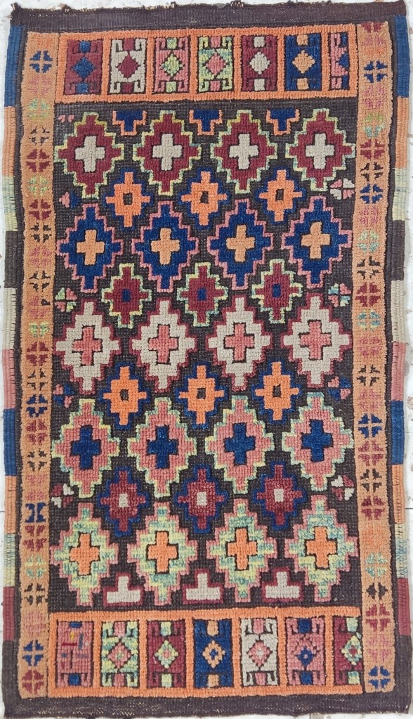Small Anatolian tribal rug (Yastik) with repeated cross-shaped motifs in red, blue, ivory, and orange on a dark ground, bordered by multi-panel tribal designs.